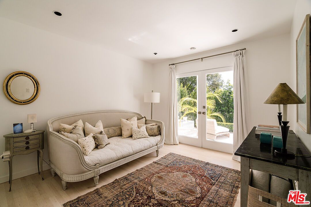 This is an interior shot of a living room featuring a daybed with decorative pillows, a vintage-style rug, and a small side table with a mirror above it. French doors lead to an outdoor area, providing natural light and a view of the greenery. The room has a bright and airy feel, with a neutral color palette and classic design elements.