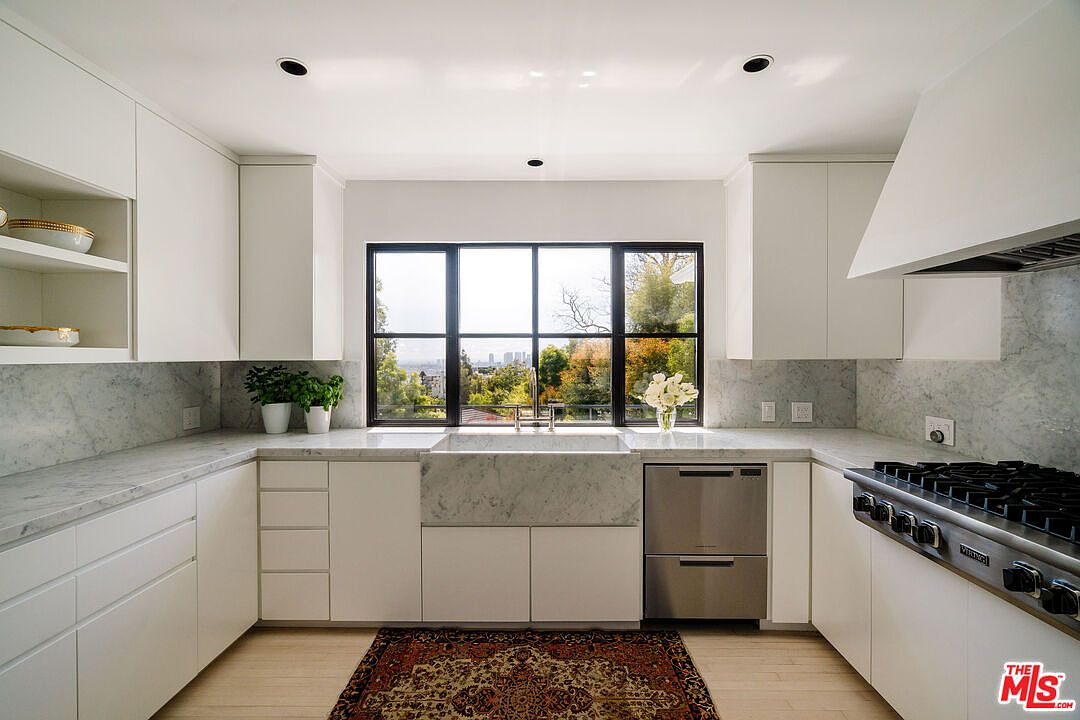 This is a bright and modern kitchen featuring white cabinetry, marble countertops and backsplash, and stainless steel appliances. A large window above the sink provides natural light and a view of the outdoors. The kitchen is clean and well-organized, creating a welcoming and functional space.
