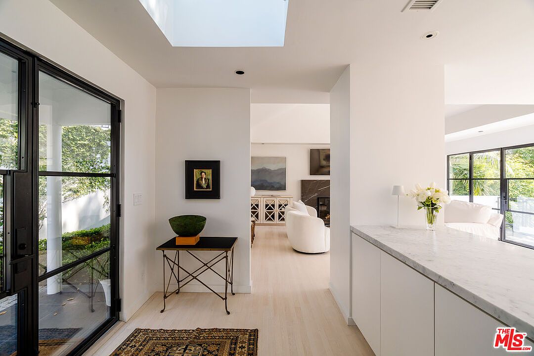 This interior shot showcases a bright and modern hallway with light wood flooring and white walls. A black-framed glass door leads to the exterior, while an opening reveals a living area with a white sofa and artwork. The space is well-lit with a skylight and recessed lighting, creating a clean and inviting atmosphere.
