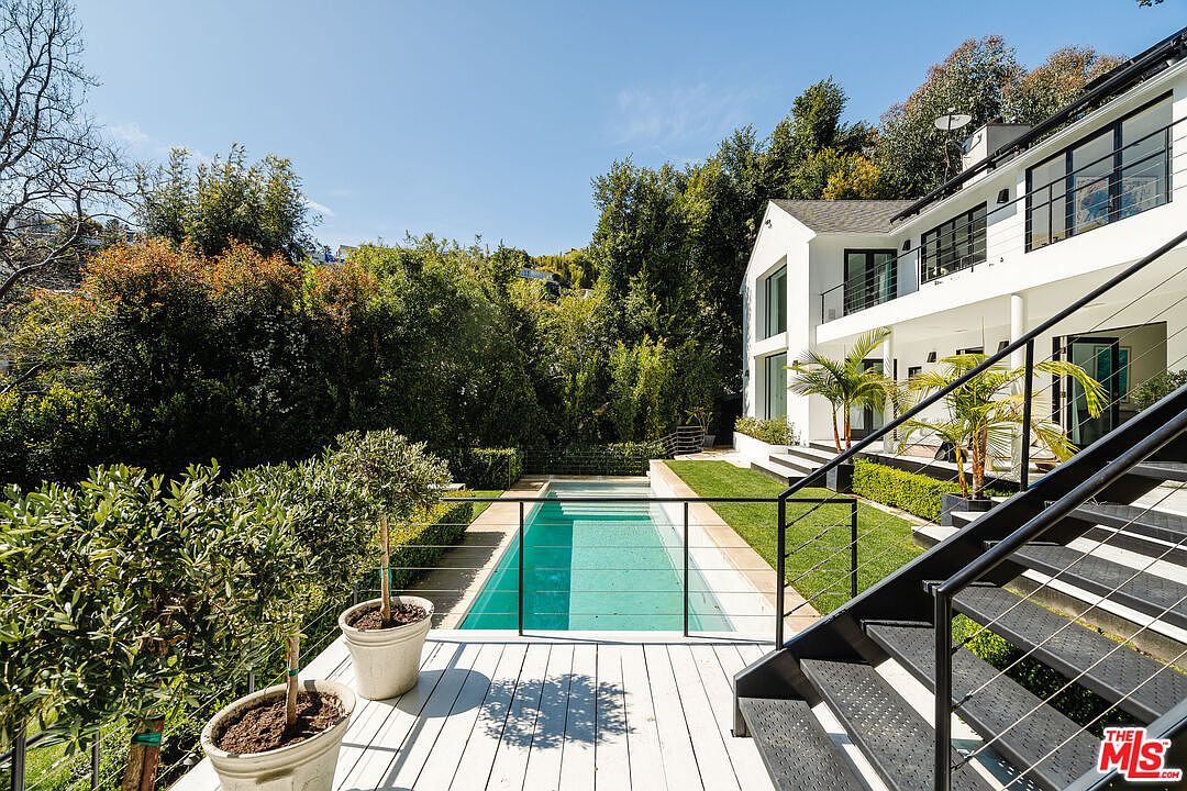This image showcases a modern home's exterior with a focus on the pool and surrounding landscape. The pool is rectangular and surrounded by a white deck and lush greenery, creating a private and serene atmosphere. A modern white house with black framed windows is visible in the background, complemented by black metal stairs leading to the deck, enhancing the property's contemporary appeal.