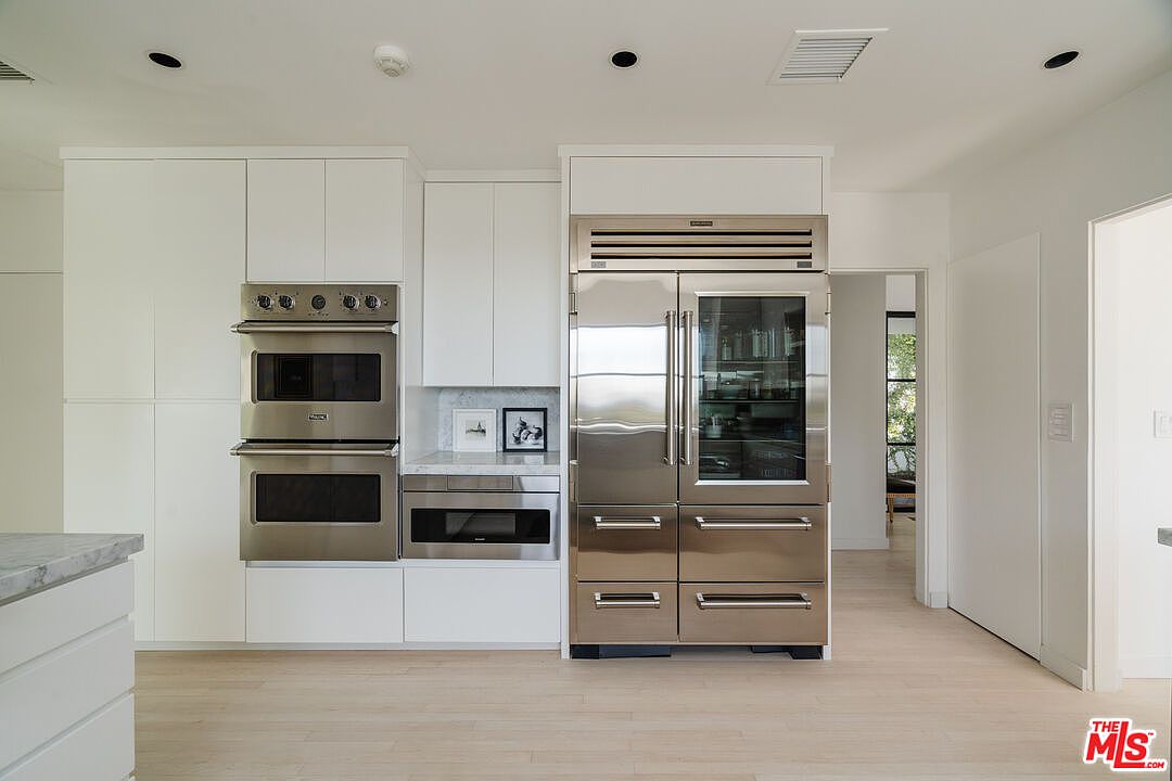 This is a well-lit kitchen featuring white cabinetry and stainless steel appliances. The appliances include a double oven, microwave, and a large refrigerator with a glass door. The kitchen has a clean, modern aesthetic with light wood flooring and a glimpse into an adjacent room.