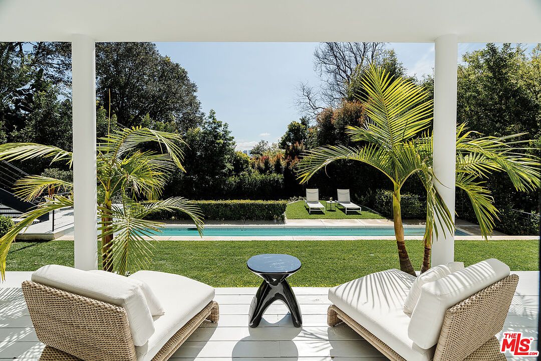 This image showcases a luxurious outdoor patio or balcony area, featuring two lounge chairs with white cushions and a small black table. The space overlooks a well-manicured lawn and a sparkling pool, framed by lush greenery and palm trees. The view is captured from a covered area with white pillars, creating a sense of privacy and relaxation.