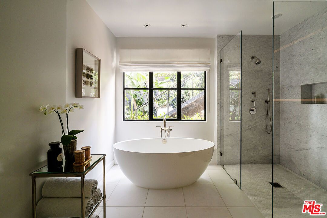 This is a bright and modern primary bathroom featuring a freestanding white bathtub positioned in front of a large window with black frames. A glass-enclosed shower with marble walls and modern fixtures is located to the right, while a stylish side table with towels and decorative items sits to the left. The overall impression is clean, luxurious, and spa-like.