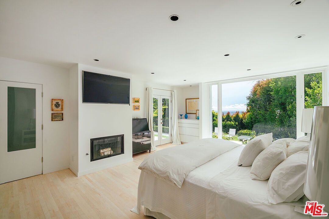This is a bright and airy primary bedroom featuring a large bed with white linens, a modern fireplace with a mounted television above, and floor-to-ceiling windows offering a view of the lush greenery outside. The room has light wood flooring and white walls, creating a clean and serene atmosphere. A glass door leads to the outside.