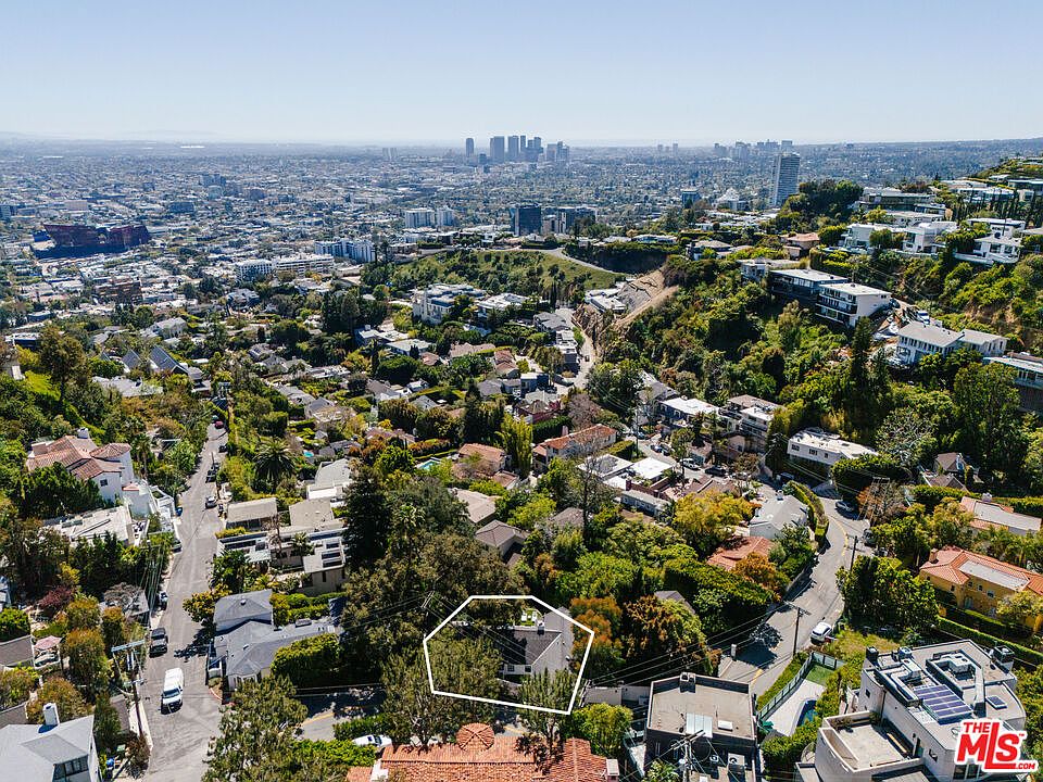 This aerial shot showcases a property nestled among lush greenery in a hillside neighborhood, with a clear view of the city skyline in the distance. The house, outlined in white, appears well-maintained and blends seamlessly with the surrounding landscape. The image emphasizes the property's desirable location and potential for privacy.
