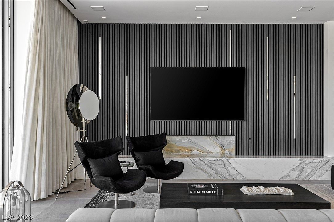 This sophisticated living room features a striking accent wall with vertical slats and integrated linear lighting flanking a wall-mounted television. Below the television, a low-profile stone hearth extends across the wall, complemented by two modern black swivel chairs and a dark coffee table. To the left, floor-to-ceiling curtains frame a large window, while a unique, oversized studio-style floor lamp adds a dramatic, artistic touch to the space.