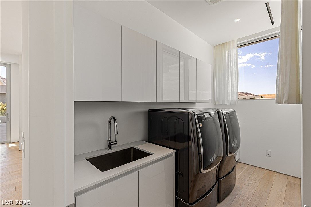 This modern laundry room features sleek, high-gloss white cabinetry and a minimalist sink area with a light-toned countertop. A pair of dark, high-capacity laundry appliances sits adjacent to the sink, while a large window with sheer curtains allows natural light to brighten the space. The room is finished with light wood-toned flooring, creating a clean and functional aesthetic.