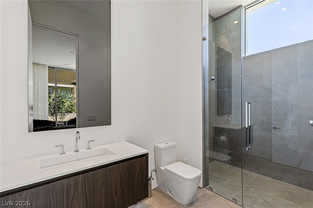 This modern bathroom features a sleek, dark wood vanity with a white countertop and a rectangular integrated sink. A large, frameless mirror reflects the adjacent room, while a glass-enclosed walk-in shower with neutral-toned wall tiles and a high window adds to the contemporary aesthetic. The space is finished with a white toilet and light-colored flooring, creating a clean and minimalist atmosphere.