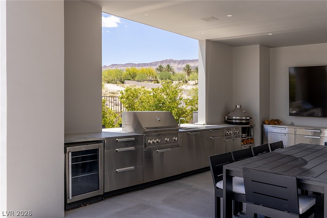 This covered outdoor kitchen and dining area features sleek stainless steel cabinetry, a built-in grill, and a beverage refrigerator. A large dining table sits adjacent to the cooking station, while the open-air design provides a seamless transition to the desert landscape and mountain views beyond. The space is modern and functional, perfect for alfresco entertaining.