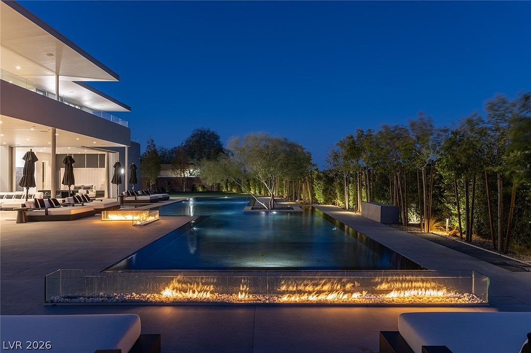 This stunning evening view captures a modern luxury backyard featuring a long, rectangular swimming pool as the central focal point. The scene is illuminated by warm, linear fire features set in glass enclosures and soft landscape lighting that highlights the surrounding trees and manicured greenery. The perspective from the patio showcases a seamless transition between the indoor living space and the outdoor lounge area, creating an inviting and sophisticated atmosphere.