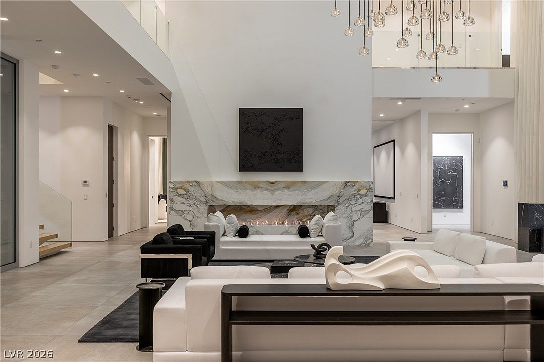 This expansive, double-height living room features a minimalist aesthetic with a prominent stone-clad chimney breast housing a linear fireplace. The space is furnished with white sofas and black accent chairs, arranged around a central seating area, while a sculptural light fixture hangs from the high ceiling. The open-concept layout flows into adjacent hallways, showcasing a clean, modern design with neutral tones and high-end finishes.
