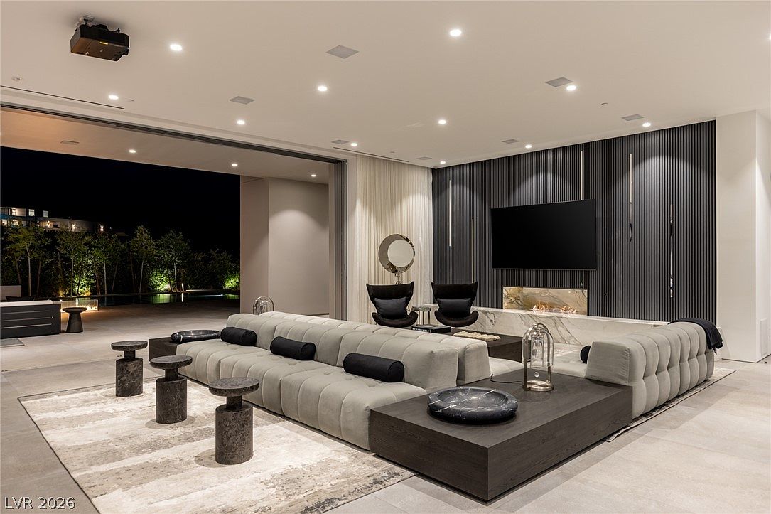 This expansive, contemporary living room features a large, modular sectional sofa arranged around a low-profile coffee table. A prominent feature is the linear fireplace set into a stone-clad wall, complemented by dark, vertical slat paneling that adds texture to the space. Large, retractable glass walls open the room to an outdoor patio, creating a seamless transition between indoor and outdoor living areas.