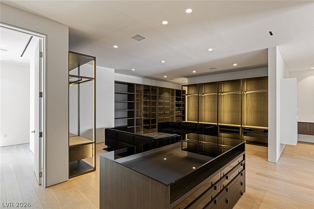 This expansive, modern walk-in closet features a central island with a dark finish and a glass top, surrounded by extensive custom cabinetry and open shelving. The room is illuminated by recessed lighting and integrated cabinet lights, creating a sophisticated and organized atmosphere. Light wood flooring runs throughout the space, which also includes a dark-toned sofa for seating.