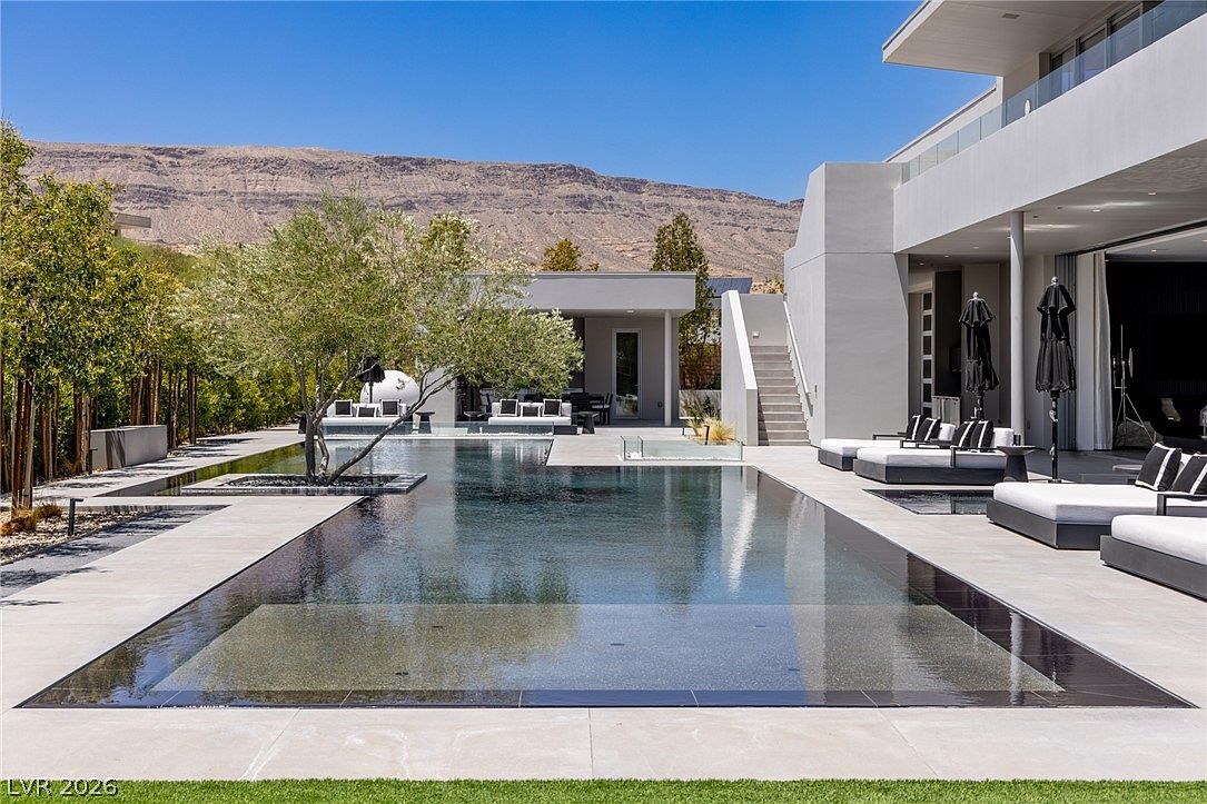 This luxurious outdoor space features a sleek, rectangular infinity-edge pool as its centerpiece, set against a backdrop of arid mountains. The modern architecture includes a covered patio area with lounge seating, clean lines, and a staircase leading to an upper level. The scene is bathed in bright, clear daylight, emphasizing the minimalist design and the seamless transition between the indoor and outdoor living areas.