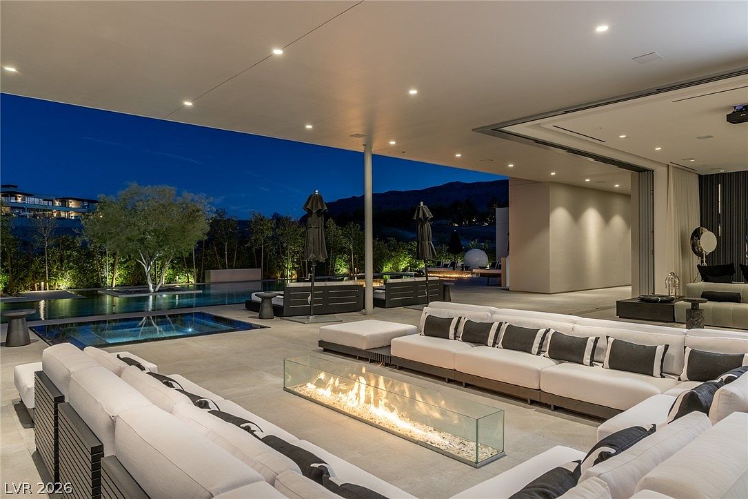 This expansive covered patio offers a seamless transition between indoor and outdoor living, featuring a large, modern sectional sofa arranged around a rectangular glass-enclosed fire feature. The space overlooks a sleek infinity-edge pool and a landscaped backyard, all set against a backdrop of distant mountains at twilight. The design emphasizes clean lines, contemporary luxury, and an open-air atmosphere perfect for evening entertaining.