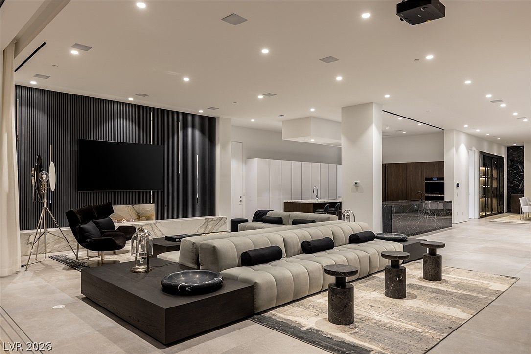 This expansive, open-concept living area features a contemporary design with a large, modular sectional sofa centered on a patterned area rug. A striking dark-paneled wall serves as a focal point, incorporating a television and a sleek, low-profile stone fireplace surround. The space flows seamlessly into a modern kitchen area in the background, characterized by clean lines, neutral tones, and recessed lighting throughout the ceiling.