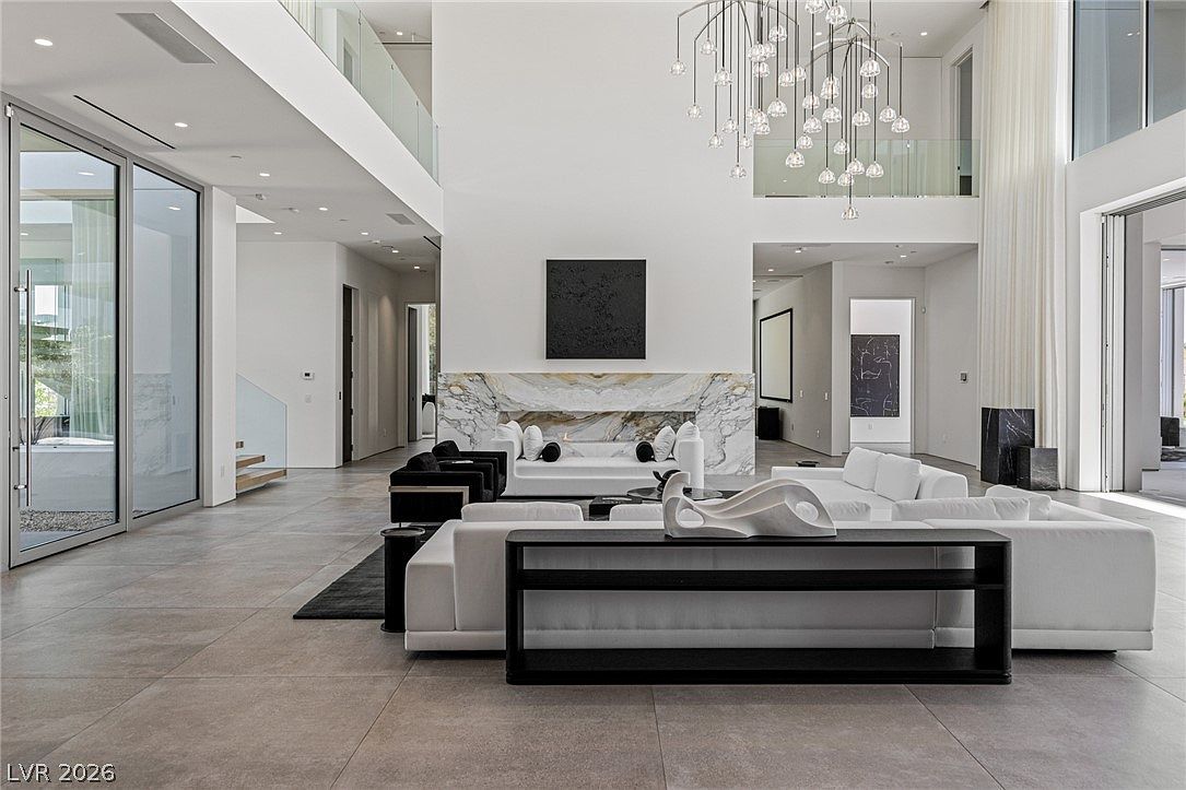 This expansive, double-height living room features a minimalist aesthetic with a neutral color palette and large-format tile flooring. A prominent stone-clad chimney breast serves as the focal point, flanked by white sofas and a dark console table in the foreground. The space is illuminated by a dramatic, multi-pendant chandelier and benefits from abundant natural light provided by large glass sliding doors.