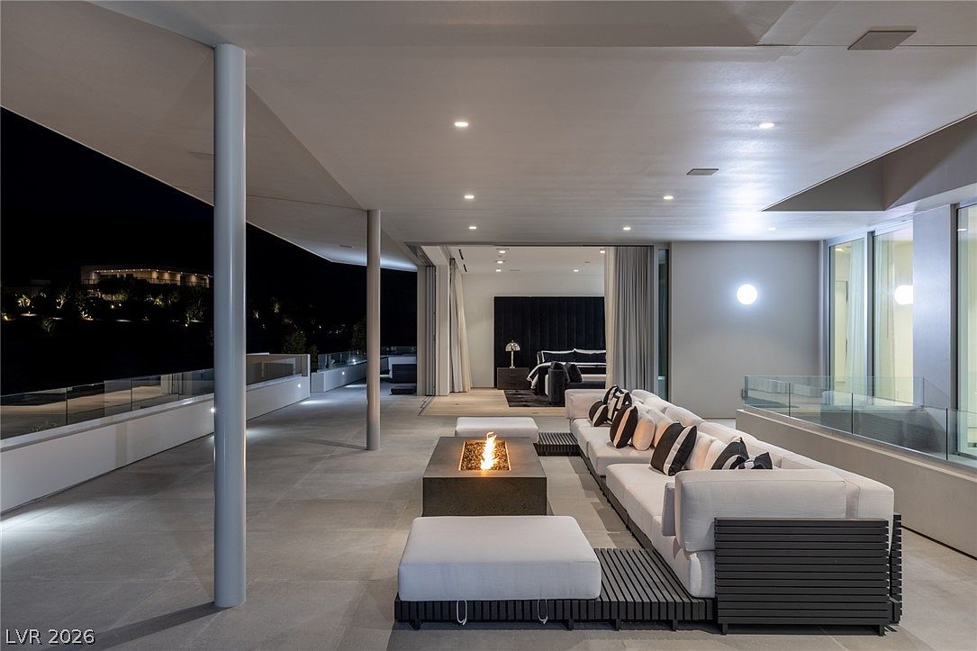 This expansive, modern covered patio offers a seamless transition from the indoor bedroom to an outdoor lounge area. The space features a large, low-profile sectional sofa arranged around a central rectangular fire pit, set against a backdrop of glass railings and a dark night sky. The clean lines, neutral color palette, and recessed ceiling lighting create a sophisticated and tranquil atmosphere perfect for evening relaxation.