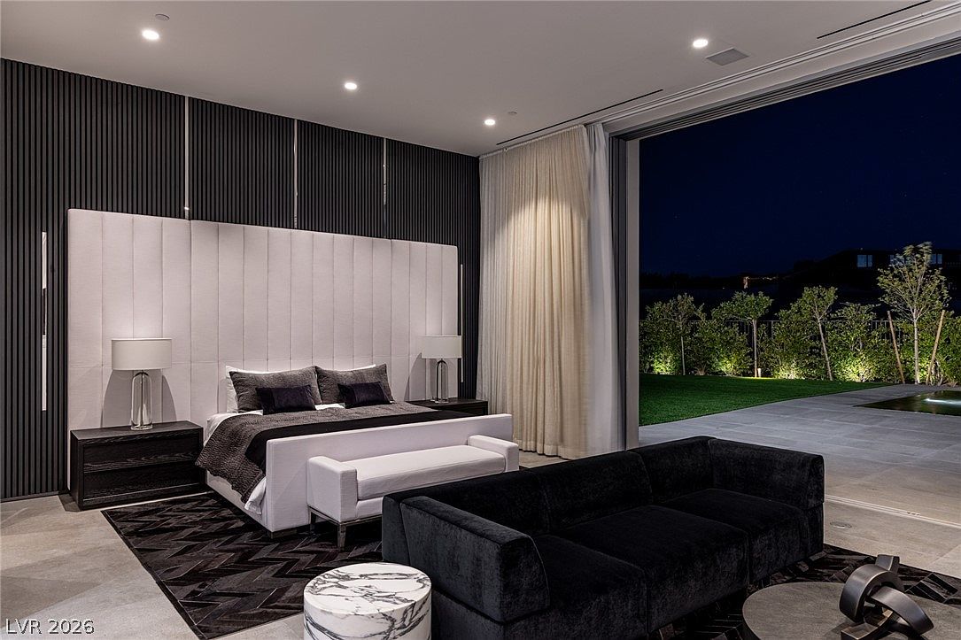 This luxurious primary bedroom features a large bed with a tall, upholstered headboard set against a dark, textured accent wall. A sleek black sofa sits in the foreground, while expansive floor-to-ceiling glass doors open to a private outdoor patio, seamlessly blending indoor and outdoor living. The room is illuminated by recessed lighting and elegant bedside lamps, creating a sophisticated and tranquil atmosphere.