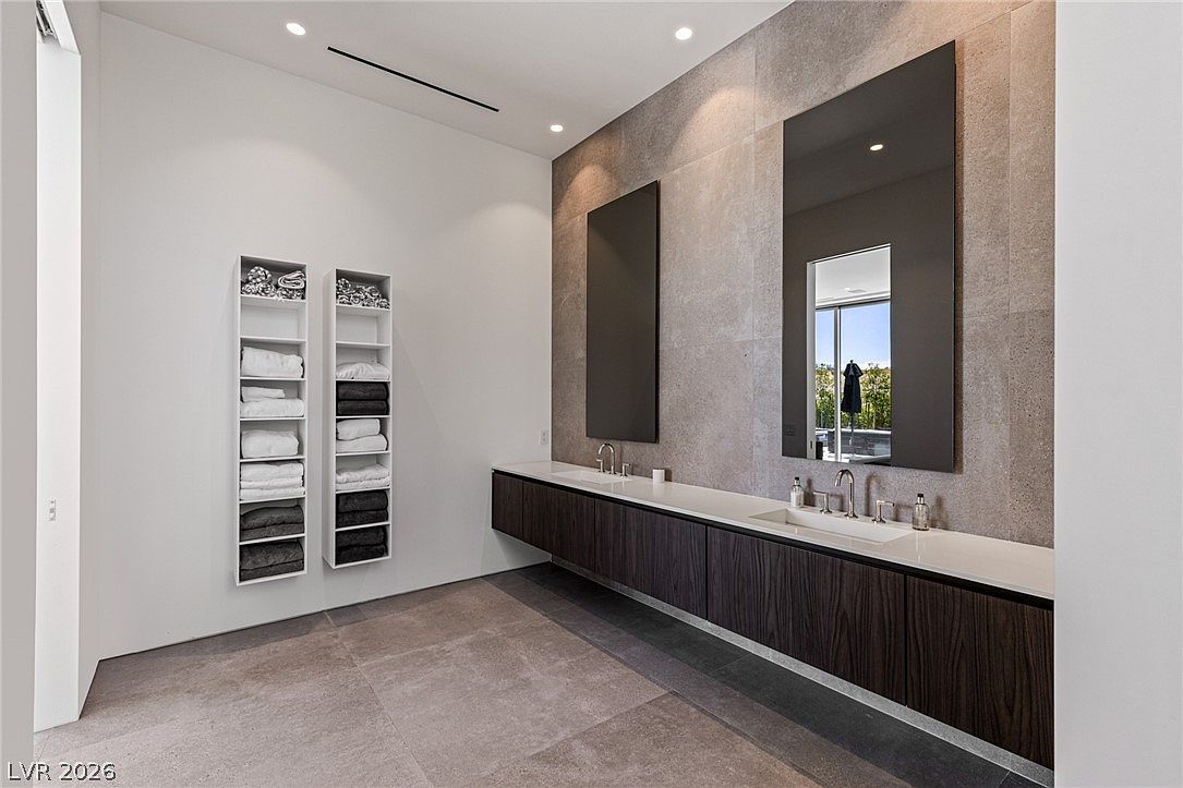 This modern primary bathroom features a long, floating vanity with dark wood cabinetry and a sleek white countertop. Two rectangular mirrors are mounted against a large, textured stone accent wall, complemented by minimalist fixtures and recessed lighting. The space is finished with large-format floor tiles and includes built-in shelving units stocked with neatly folded towels.