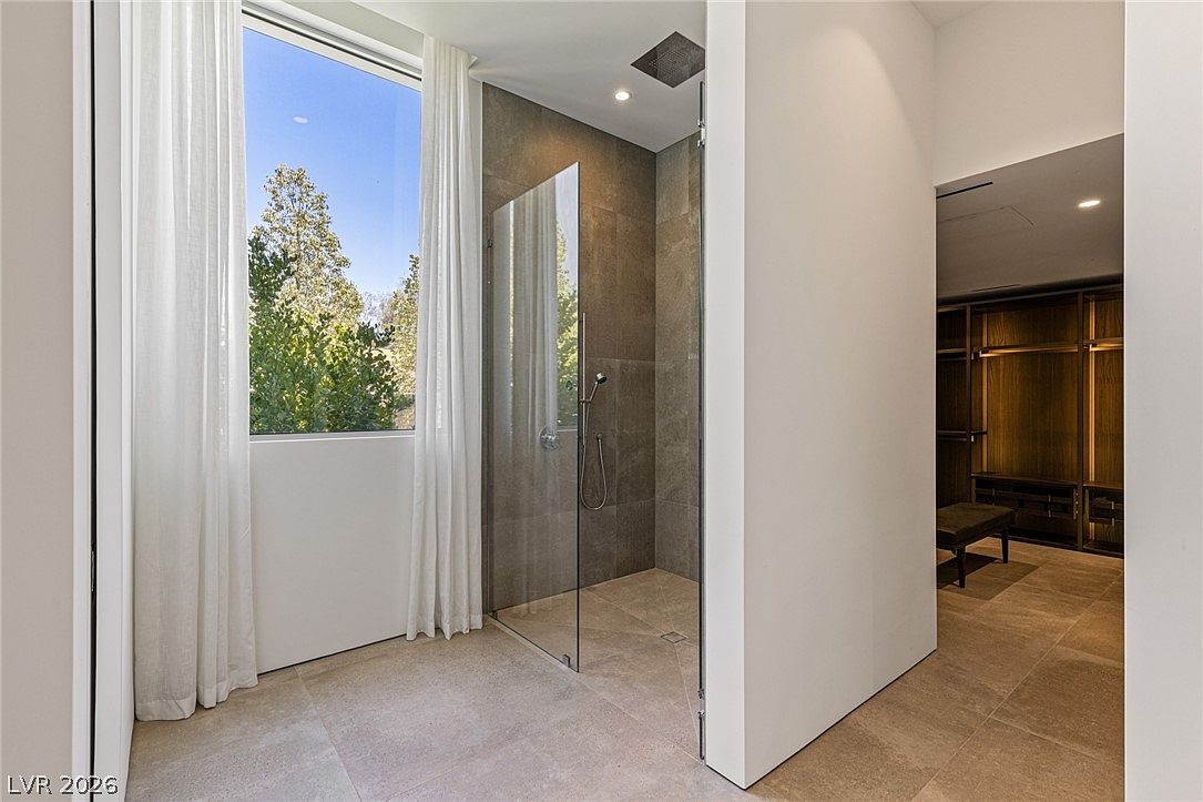 This modern bathroom features a spacious walk-in shower with large-format stone tiles and a glass enclosure. A large window with sheer curtains allows natural light to fill the space, while an adjacent doorway leads into a walk-in closet with built-in shelving. The neutral color palette and clean lines create a serene and contemporary atmosphere.