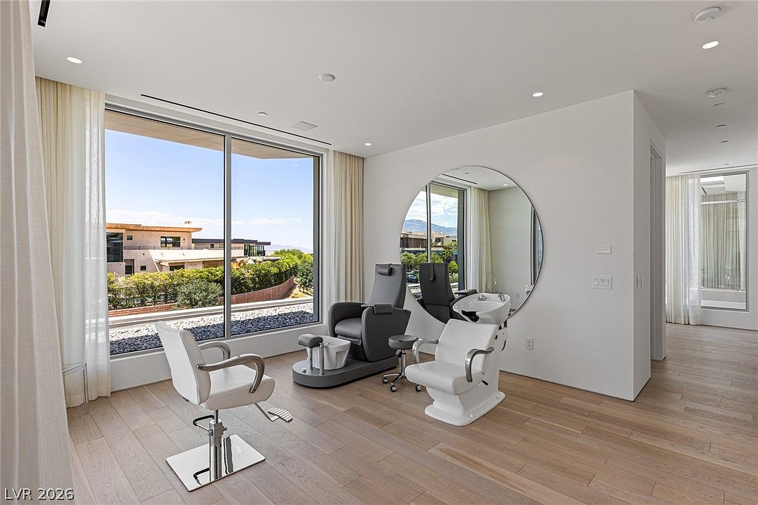 This image showcases a private salon or spa room within a home, featuring a professional styling chair, a pedicure station, and a shampoo bowl. The space is bright and airy, highlighted by large windows that offer a scenic view of the surrounding neighborhood and mountains. A large, circular mirror serves as a focal point on the wall, while light wood flooring and neutral tones create a clean, modern aesthetic.