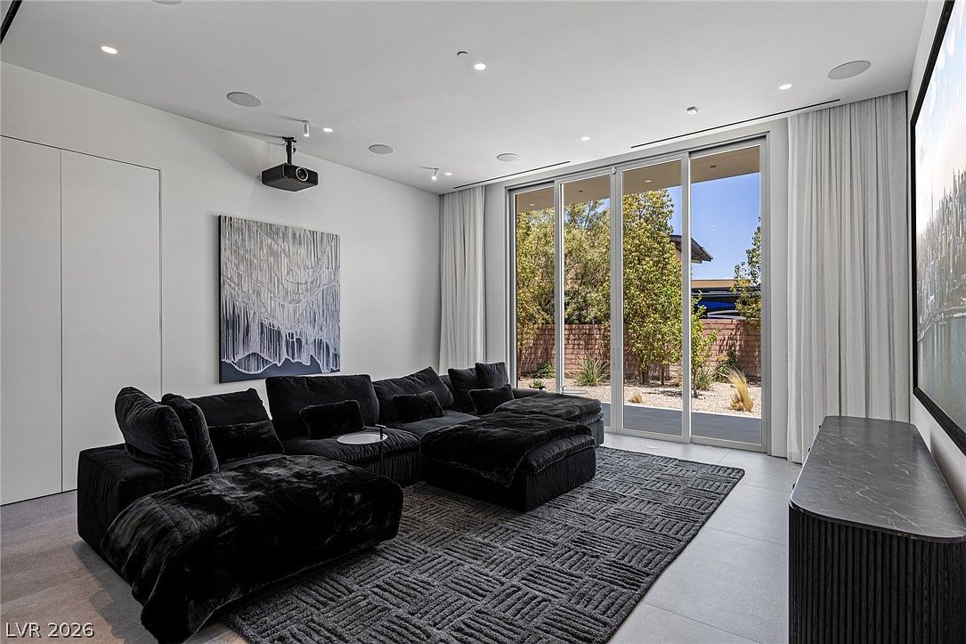 This modern media room features a large, plush black sectional sofa centered on a textured geometric rug. A projector mounted on the ceiling faces a large screen on the right wall, while floor-to-ceiling sliding glass doors provide abundant natural light and a view of the outdoor landscaping. The minimalist aesthetic is complemented by neutral walls, recessed lighting, and a sleek dark console table.