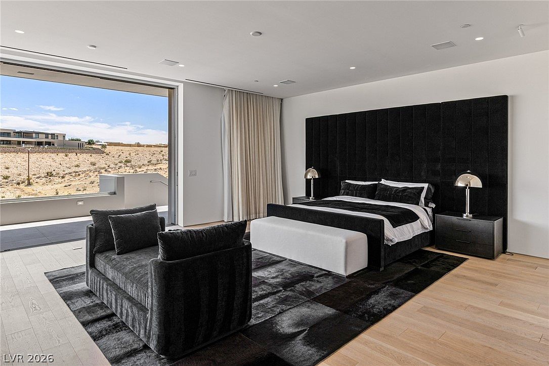 This spacious primary bedroom features a modern, minimalist aesthetic with a striking black velvet-upholstered headboard wall and a matching dark-toned area rug. A plush white bench sits at the foot of the bed, while a contemporary chaise lounge is positioned to face the large, sliding glass doors that open to an expansive desert view. The room is accented by light wood flooring and sleek, metallic bedside lamps, creating a sophisticated and serene atmosphere.