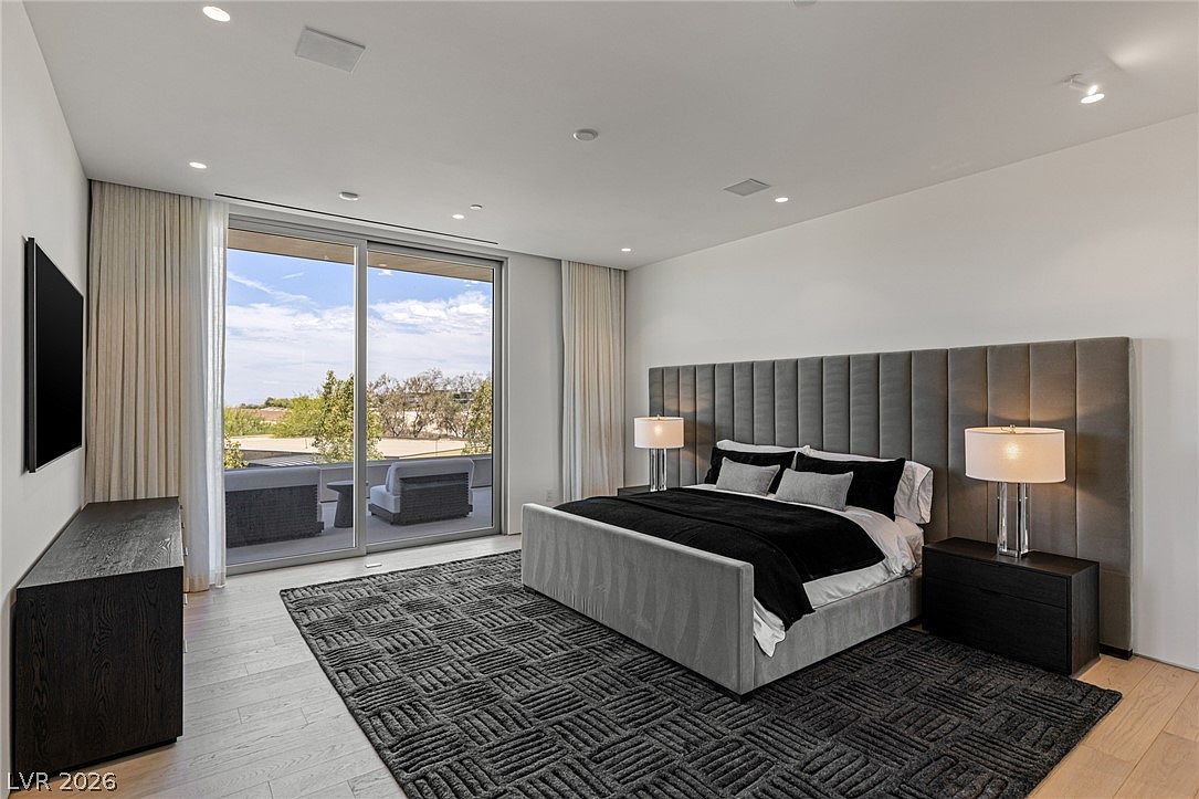 This spacious primary bedroom features a contemporary design with a large, upholstered headboard and a textured, geometric-patterned area rug. The room is anchored by a platform bed and flanked by two modern bedside tables with minimalist lamps, while a sleek dark wood dresser sits opposite the bed. Large sliding glass doors provide access to a private balcony, allowing natural light to fill the room and offering an expansive view of the outdoors.