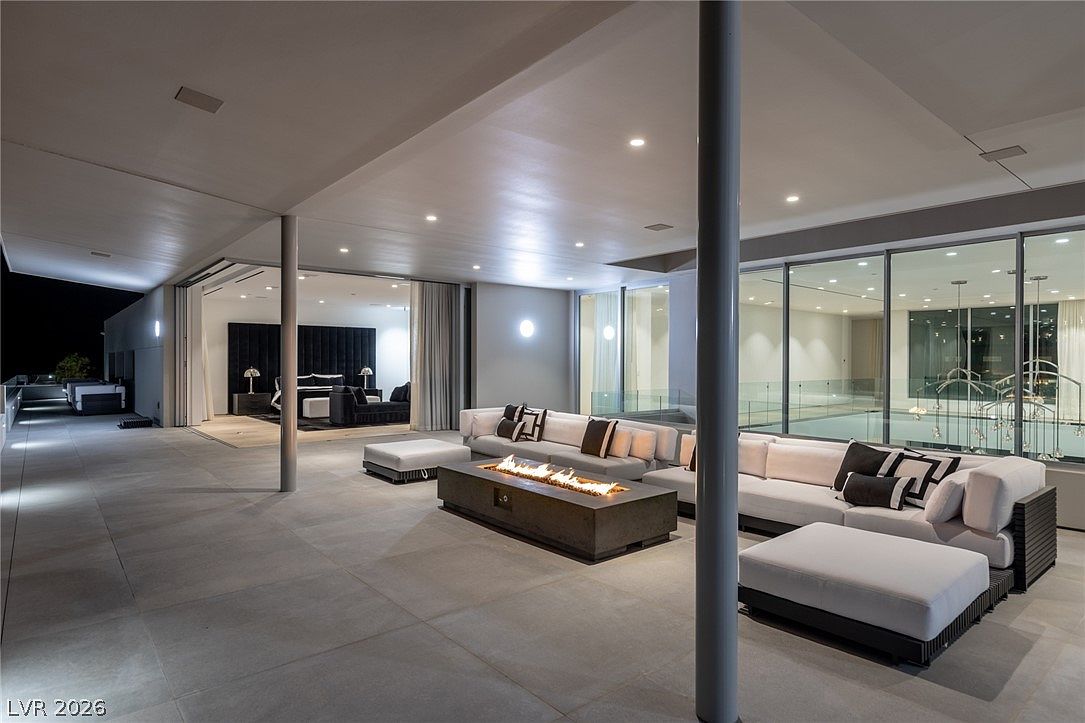This expansive, modern covered patio offers a seamless transition between indoor and outdoor living, featuring large glass walls that open into a bedroom suite. The space is anchored by a rectangular fire pit surrounded by plush, neutral-toned sectional seating, creating an inviting atmosphere for evening relaxation. The clean lines, recessed lighting, and polished tile flooring contribute to a sophisticated, high-end aesthetic overlooking a glass-railed terrace.