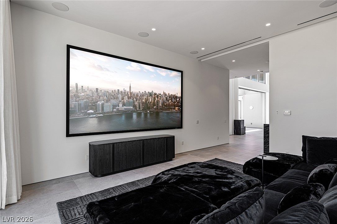 This modern home theater or media room features a large, wall-mounted screen displaying a cityscape, positioned above a sleek, dark, ribbed media console. A plush, dark-toned sectional sofa occupies the foreground, set upon a textured rug that complements the neutral tile flooring. The room transitions into an open hallway, showcasing a minimalist aesthetic with clean white walls and recessed lighting.