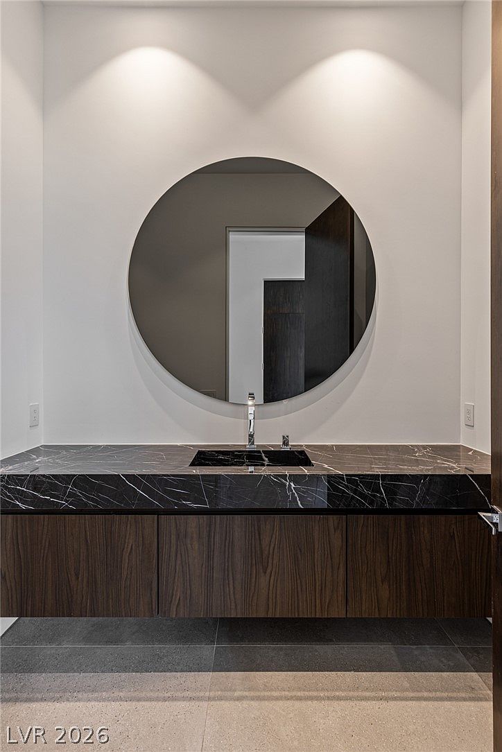 This modern powder room features a floating vanity with a dark wood-toned finish and a striking black stone countertop with white veining. A large, circular mirror is centered above the integrated sink, complemented by minimalist wall-mounted fixtures. The space is finished with neutral walls and dark floor tiles, creating a sophisticated and contemporary aesthetic.