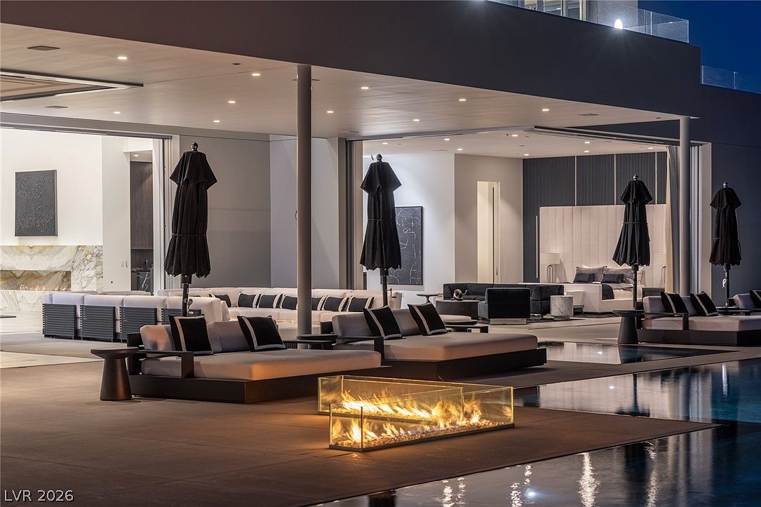 This luxurious outdoor living space features a seamless transition between the indoor bedroom area and a sprawling covered patio. The scene is anchored by a sleek, rectangular swimming pool with a modern glass-enclosed fire feature at its edge. Multiple lounge areas with contemporary dark-toned furniture and black umbrellas create a sophisticated, resort-like atmosphere perfect for evening entertaining.