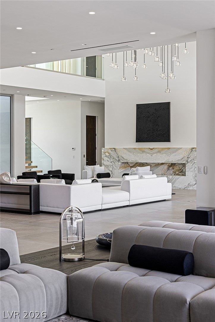 This expansive, open-concept living area features a double-height ceiling and a sophisticated, modern aesthetic. A large, white modular sofa arrangement serves as the centerpiece, complemented by a striking stone-clad fireplace surround and a minimalist black art piece. The perspective captures the vastness of the room, highlighting the elegant pendant lighting and the seamless transition to the staircase in the background.