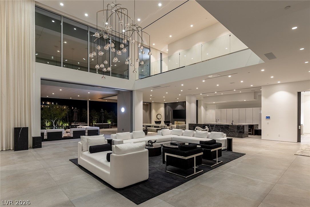 This expansive, double-height living room features a contemporary open-concept design with a large white sectional sofa and black accent chairs arranged on a dark area rug. A striking, multi-tiered chandelier hangs from the high ceiling, while large glass walls provide a seamless transition to the outdoor patio and pool area. The space is characterized by its minimalist aesthetic, neutral color palette, and a view into a sleek kitchen with a dark stone island in the background.