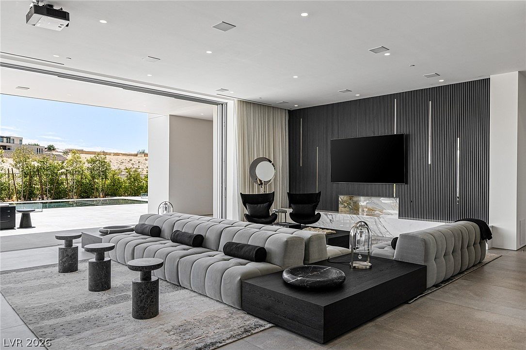 This expansive, modern living room features a large, modular grey sofa set against a striking dark-slatted accent wall with a mounted television. A tall, light-toned stone fireplace surround adds texture to the room, while floor-to-ceiling glass doors slide open to reveal a seamless transition to the outdoor pool area. The space is defined by its minimalist aesthetic, neutral color palette, and high-end contemporary furnishings.