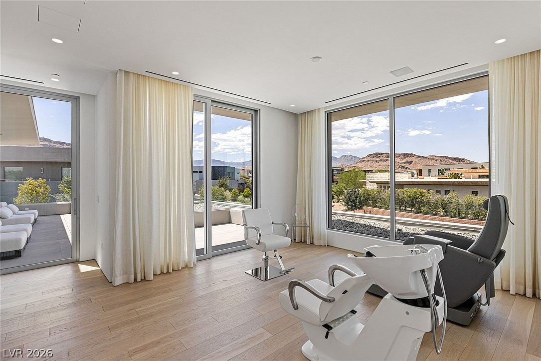 This image depicts a professional-style salon or spa room located within a residence, featuring a reclining shampoo station and a styling chair. The space is bright and airy, characterized by light wood flooring and expansive floor-to-ceiling windows that provide panoramic views of the surrounding desert landscape. A sliding glass door leads out to an adjacent patio, enhancing the indoor-outdoor flow of the room.