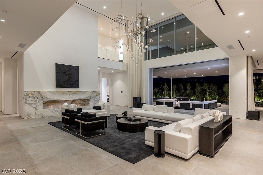 This expansive, double-height living room features a minimalist aesthetic with large, light-colored floor tiles and a dark area rug anchoring the seating area. A striking, multi-tiered chandelier serves as a focal point, while a long, linear fireplace is set into a stone-clad wall. The space seamlessly transitions to an outdoor patio through massive, retractable glass walls, creating an open-concept indoor-outdoor living experience.