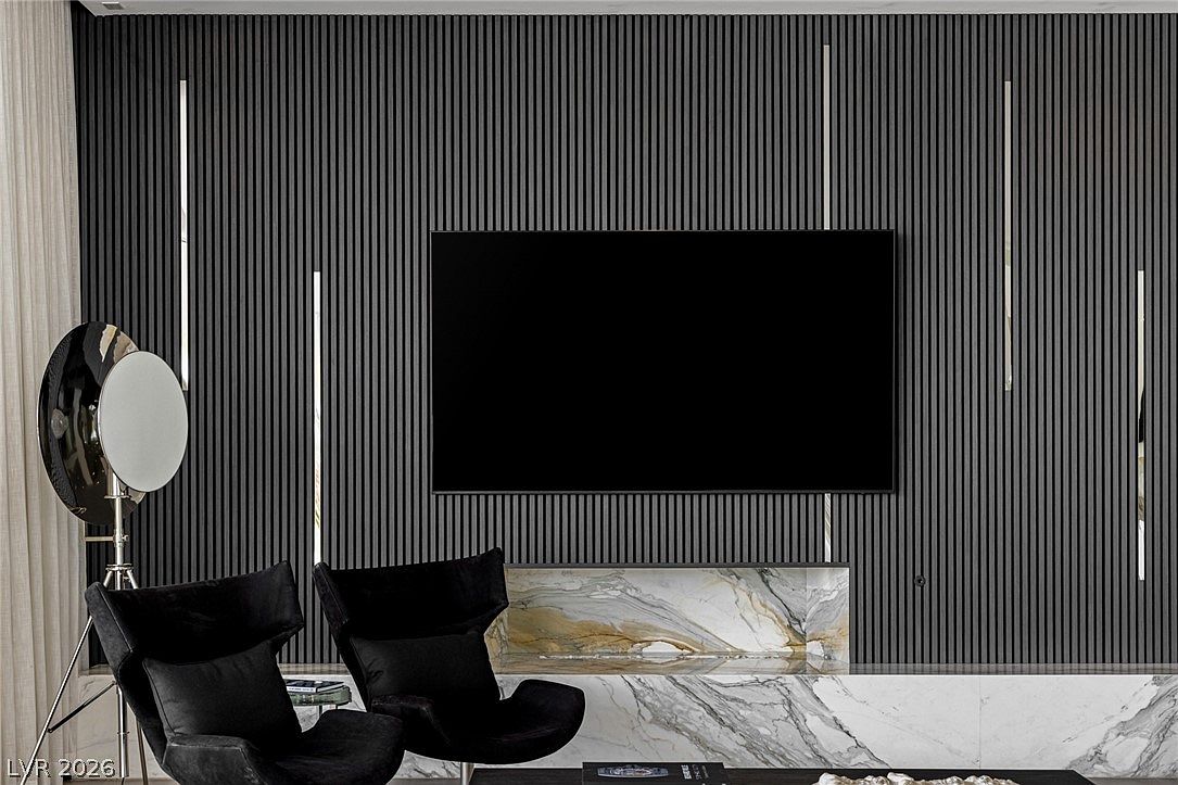 This sophisticated living room features a striking accent wall with vertical slats and integrated linear lighting flanking a large wall-mounted television. Below the television, a stone-clad hearth with dramatic veining provides a luxurious focal point. Two modern, dark-upholstered lounge chairs are positioned in the foreground, complemented by a unique, circular floor lamp that adds a sculptural element to the space.