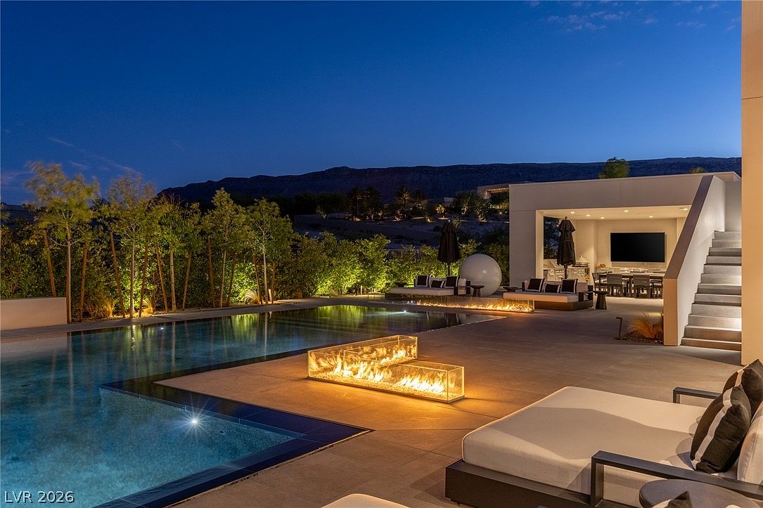 This luxurious outdoor living space features a sleek, rectangular swimming pool as the centerpiece, complemented by a modern fire feature set in a glass enclosure on the patio. The area includes a covered lounge space with comfortable seating and a dining area, all set against a backdrop of desert landscaping and distant mountains at dusk. The scene is illuminated by warm, ambient landscape lighting, creating an inviting and sophisticated atmosphere for evening relaxation.