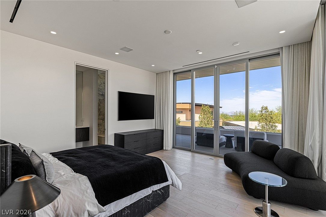 This contemporary primary bedroom features light wood flooring and a minimalist aesthetic with a black bed frame and matching dresser. A large, curved sofa sits near expansive glass sliding doors that provide access to a private balcony and offer views of the surrounding landscape. The room is illuminated by recessed lighting and natural light, creating a bright and airy atmosphere.