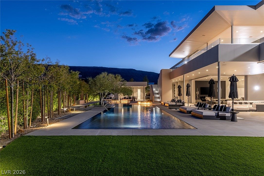 This stunning contemporary estate features a sleek, rectangular infinity-edge pool as the centerpiece of a meticulously landscaped backyard at dusk. The expansive patio area is furnished with modern lounge seating and umbrellas, seamlessly connecting to the home's indoor living space through large glass walls. The scene is illuminated by warm ambient lighting, highlighting the clean architectural lines and the lush bamboo border against the backdrop of a mountain silhouette.