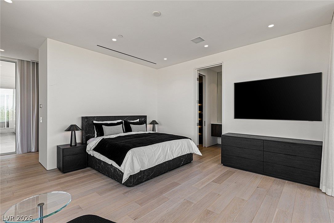 This spacious primary bedroom features a modern aesthetic with light wood flooring and crisp white walls. A large bed with a dark, textured headboard serves as the focal point, flanked by matching nightstands and lamps, while a wall-mounted television sits above a long, dark dresser. To the left, large glass doors provide access to an outdoor space, and a doorway on the right leads into a walk-in closet area.