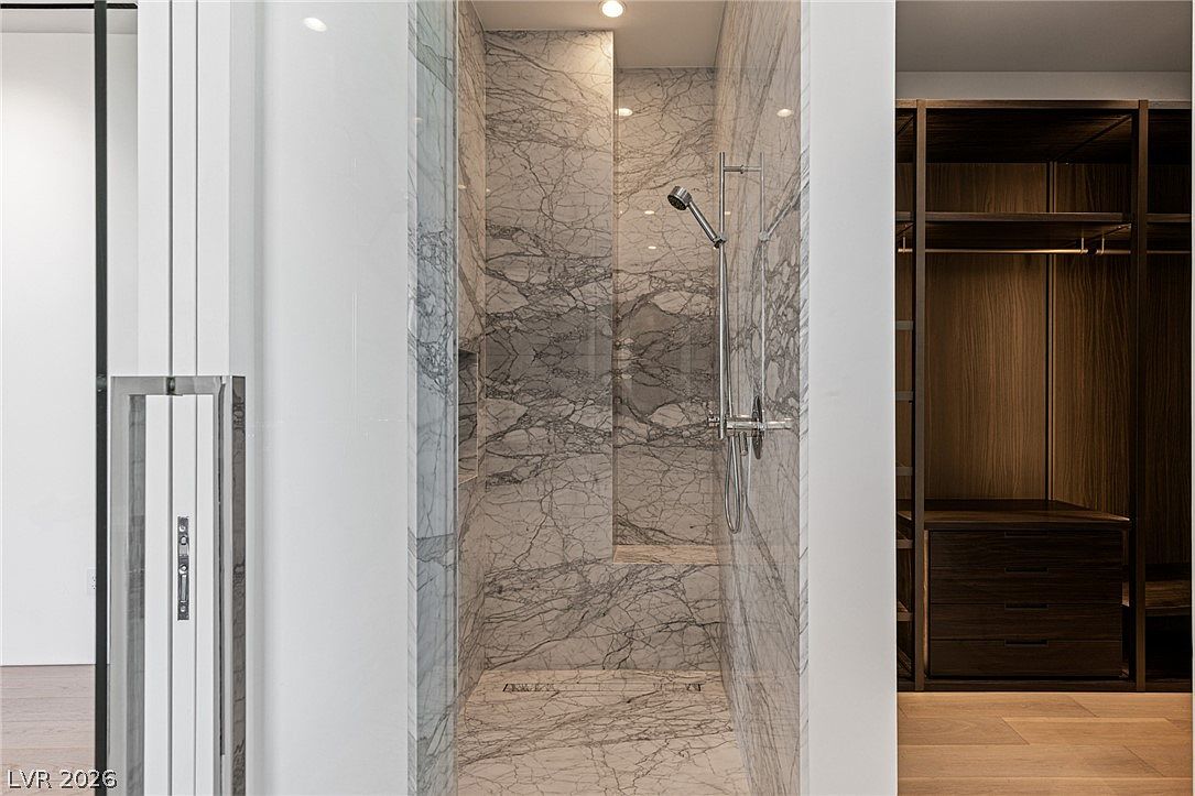 This image showcases a luxurious walk-in shower featuring extensive stone tiling with a prominent veined pattern. The shower includes a built-in bench and a modern handheld shower fixture, positioned adjacent to a walk-in closet with dark wood shelving. The perspective captures the transition between the tiled shower area and the adjacent room, highlighting a high-end, contemporary design aesthetic.