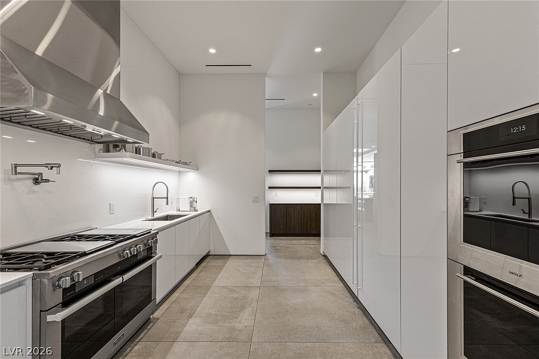 This sleek, contemporary kitchen features a galley-style layout with high-gloss white cabinetry and professional-grade stainless steel appliances, including a Wolf oven. The space is defined by large-format neutral floor tiles and a minimalist aesthetic, with a long, clean countertop and a wall-mounted faucet. A transition area leads to a secondary space with floating shelves, creating a bright and highly organized culinary environment.