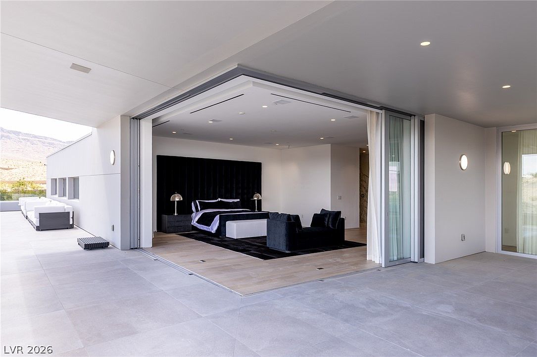 This primary bedroom features a seamless transition to an expansive outdoor patio through large, retractable glass walls that open the space to the desert landscape. The room is anchored by a dark, upholstered headboard and a matching velvet sofa, set against a neutral color palette and light wood flooring. The perspective captures the indoor-outdoor flow, highlighting the modern architectural design and the integration of the living space with the exterior environment.