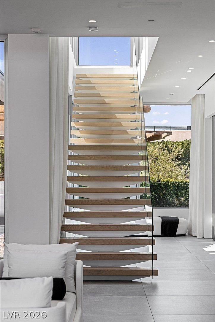 This image captures a striking, modern staircase featuring light wood treads and a minimalist glass railing, serving as a central architectural focal point. The staircase is positioned beneath a large skylight that floods the area with natural light, creating an airy and open atmosphere. The surrounding space is finished in a clean, neutral palette with large-format tile flooring and a glimpse of a lush outdoor garden through the adjacent windows.