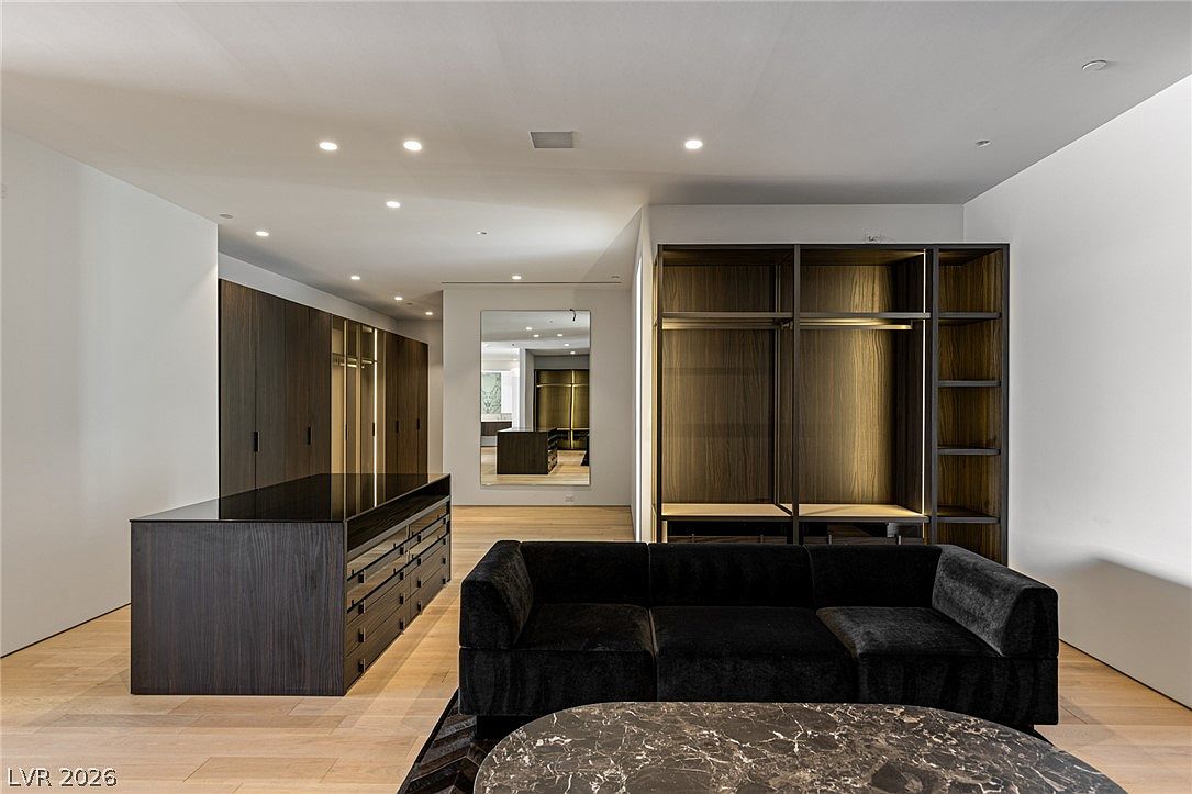 This expansive, modern walk-in closet features dark wood cabinetry, open shelving, and a central island with a dark surface and integrated drawers. A plush black sofa sits in the foreground, facing a marble-topped coffee table, while a large mirror reflects the depth of the room. The space is illuminated by recessed lighting, creating a sophisticated and organized atmosphere.