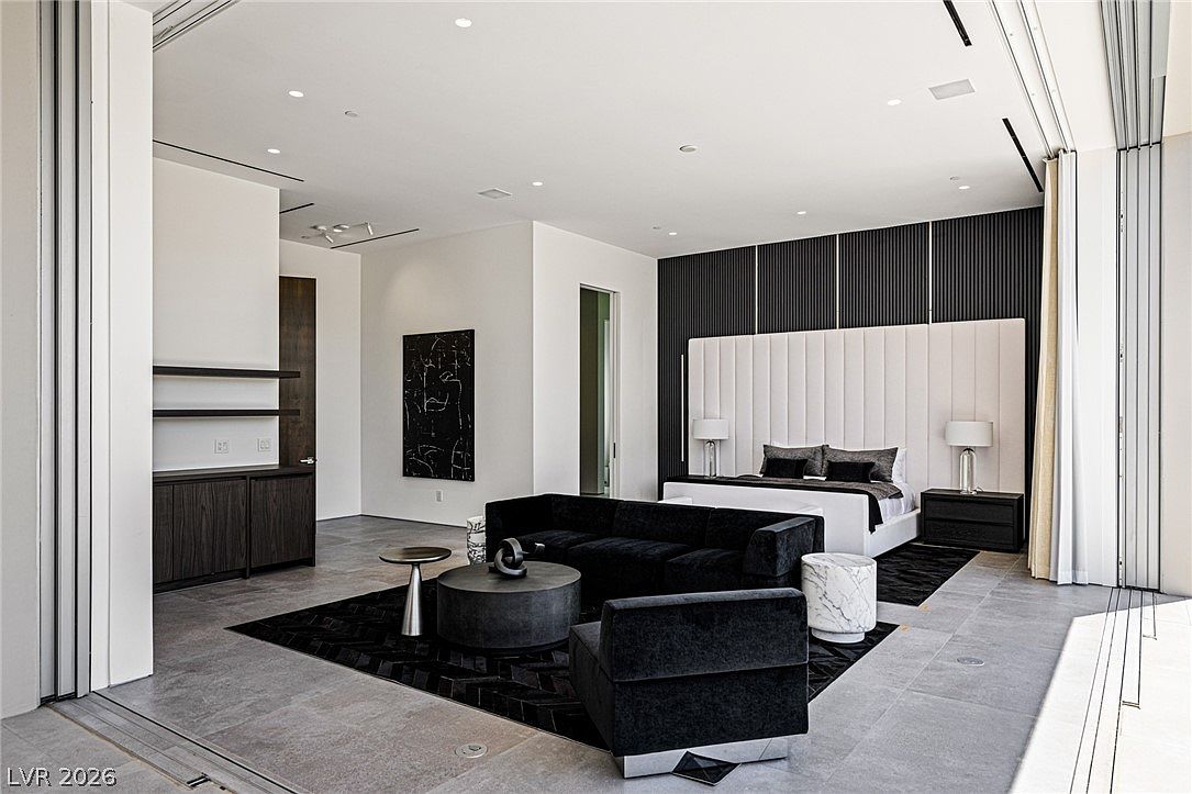 This expansive primary bedroom features a contemporary design with a neutral color palette, highlighted by a large, upholstered headboard set against a dark, vertical-slatted accent wall. The room includes a dedicated seating area with a black sofa and circular coffee table, all resting on a dark patterned rug atop large-format stone flooring. Large glass doors open to the side, creating a seamless transition to the outdoors and filling the space with natural light.