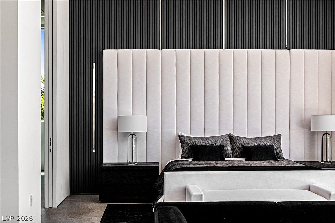 This sophisticated primary bedroom features a dramatic, oversized upholstered headboard with vertical channel tufting that spans the width of the bed. The wall behind the bed is finished with dark, vertical-slatted paneling, creating a high-contrast, modern aesthetic. Flanking the bed are minimalist dark wood nightstands topped with sleek, contemporary glass-base table lamps.